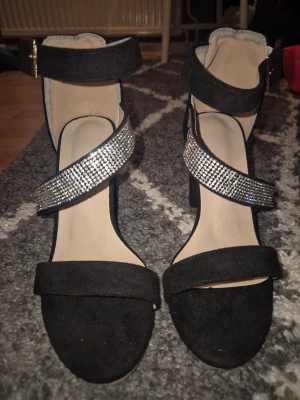Svarta sandaler med strass och klack - Stilrena svarta sandaler med bred klack och öppen tå. Remmar i mockaimitation, varav en är täckt med glittrande strass. Justerbart spänne i guld vid ankeln. Perfekta för dig som vill ha en extra touch till outfiten. Storlek 35 aldrig använda, endast testade inomhus.
