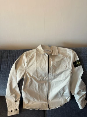 Vit overshirt med svart dragkedja - Helt ny, har använts två gånger bara !Snygg vit overshirt från Stone Island med svart dragkedja framtill och klassisk patch på vänster ärm. Overshirten har en bröstficka och knappar vid ärmsluten. Tillverkad i ett lätt material som passar perfekt till lager-på-lager-stil. 