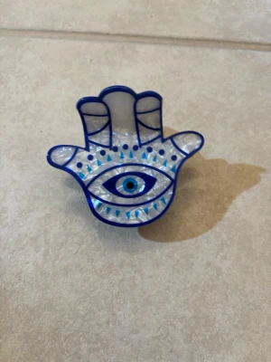 Hårklämma Hamsa med blått öga - Unik hårklämma i form av en Hamsa-hand med blått öga i mitten. Klämman är genomskinlig med blå detaljer och mönster, tillverkad i plast. Perfekt för att ge din frisyr en personlig touch och skyddande vibe.