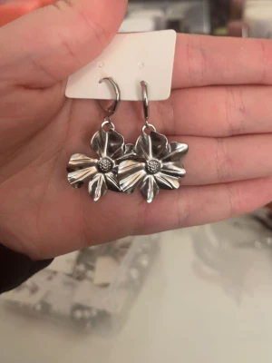 Silvriga blomformade örhängen - Säljer ett par snygga örhängen i silverfärgad metall med stora blommor som hänger ner. Blommorna har detaljerade blad och en markerad mitt, vilket ger dem en cool och unik look. Perfekta för att lyfta vilken outfit som helst!