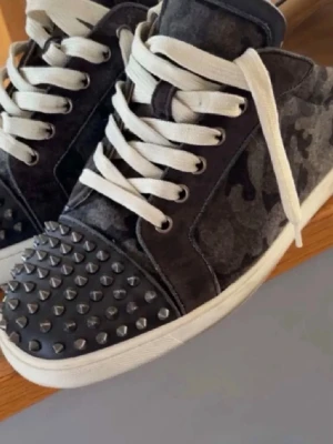 Svarta camo sneakers med nitar - Unika sneakers i svart camomönster med vita snören och vit sula. Tåpartiet är täckt av coola silverfärgade nitar och sulan har en röd undersida. Materialet ser ut att vara mocka eller textil. Perfekta för dig som vill sticka ut med din stil.