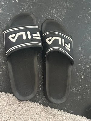Svarta Fila badtofflor - Säljer ett par svarta Fila badtofflor med vit logga och vita ränder på remmen. Klassisk slip-on modell i syntetmaterial med räfflad sula för bra grepp. Perfekta för stranden eller att chilla hemma.