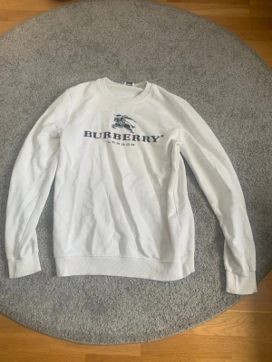 Vit sweatshirt från Burberry - Snygg vit sweatshirt från Burberry med klassiskt tryck i marinblått på bröstet och rund halsringning. Tröjan har ribbade muddar vid ärmslut och nederkant. Perfekt för en clean och stilren look.
