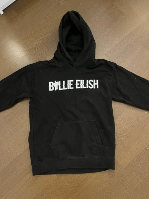 Svart Billie Eilish hoodie - Säljer en svart hoodie med vit 'Billie Eilish'-text på bröstet. Hoodien har en klassisk huva och en stor magficka framtill. Perfekt för dig som vill ha en bekväm och snygg tröja med artisttryck.