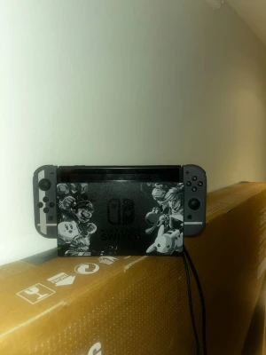 Nintendo Switch Super Smash Bros Edition - Säljer en Nintendo Switch Super Smash Bros Ultimate Edition med grå Joy-Con och specialdesignad docka med motiv från spelet. Konsolen har en svart och grå färgskala och är perfekt för gaming både hemma och på språng.