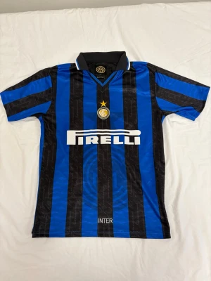 Inter Milan Fotbollströja - Säljer en klassisk Inter Milan matchtröja i blått och svart med vertikala ränder. Tröjan har korta ärmar, krage och Pirelli-logga på bröstet samt klubbmärke med stjärna. Tillverkad i lätt och ventilerande material, perfekt för fotboll eller samling. Storlek S men passar M och L utmärkt. 