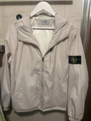 Vit vindjacka från Stone Island - Snygg vit vindjacka från Stone Island med huva och dragkedja framtill. Jackan har elastiska muddar vid ärmsluten och klassisk logotyp-patch på vänster ärm. Perfekt för blåsiga dagar och enkel att matcha med olika outfits. Aldrig använd passar m och s hör av vid frågor 