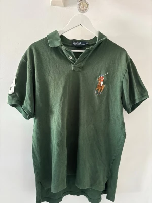 Piketröja från Ralph Lauren - Mörkgrön pikétröja från Ralph Lauren med klassisk krage och två knappar vid halsen. Stor broderad polospelare på bröstet och korta ärmar. Perfekt för en avslappnad men ändå stilren look.