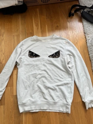 Vit sweatshirt med ögonmönster - Cool vit sweatshirt med unikt ögonmönster i svart och vitt på bröstet. Tröjan har rund halsringning och långa ärmar. Perfekt för dig som vill sticka ut med en edgy och trendig stil.