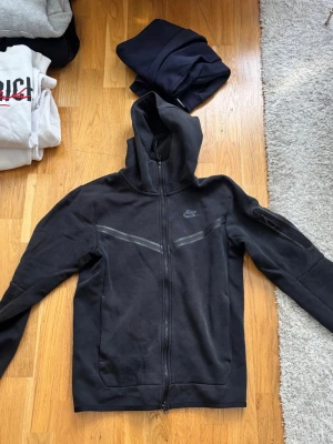 Svart Nike hoodie med dragkedja - Säljer en svart hoodie från Nike med hel dragkedja framtill och huva. Hoodien har diskreta svarta detaljer över bröstet och en ficka på ärmen. Perfekt för en sportig och avslappnad stil.