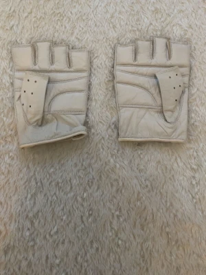 Beige fingerlösa skinnhandskar - Säljer ett par beige fingerlösa handskar i skinn med håldetaljer och öppning vid handleden. Snyggt retroinspirerat utseende, perfekta för dig som vill ha ett coolt accessoarstatement. Passar till många olika stilar.