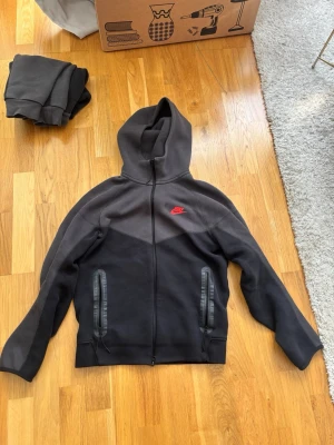 Svart Nike hoodie med dragkedja - Snygg svart hoodie från Nike med hel dragkedja framtill och två fickor med dragkedja. Hoodien har en röd Nike-logga på bröstet och en stor huva. Tillverkad i mjukt material som känns skönt mot huden. Perfekt för en avslappnad streetwear-look.