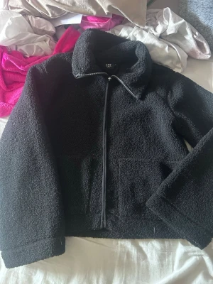 Svart teddyjacka med dragkedja - Mysig svart teddyjacka med hög krage och dragkedja framtill. Jackan har en boxy passform och är gjord i fluffigt teddy-material som håller dig varm. Perfekt för att addera en trendig touch till din outfit.