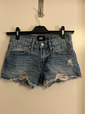 Blå denimshorts med slitningar - Säljer ett par blå jeansshorts med råa, fransiga kanter och slitna detaljer framtill. Klassisk femficksmodell med knapp och dragkedja. Perfekta för varma dagar och ger en avslappnad vibe. Använda ett fåtal gånger.