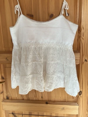 Vit spetslinnetopp med volang - Söt vit linnetopp med tunna axelband som knyts upptill. Toppen har broderade spetsdetaljer och volanger nedtill, vilket ger en romantisk känsla. Perfekt för varma dagar och somriga outfits.