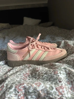 Adidas rosa Spezial sneakers - Snygga Adidas Spezial sneakers i ljusrosa mocka och läder med vita detaljer och klassiska tre ränder på sidan. Skorna har beige sula och snörning framtill. Perfekta för dig som vill ha en unik och trendig sneaker med retrovibbar.