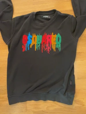Svart sweatshirt med Dsquared2-logga - Cool svart sweatshirt från Dsquared2 med ett stort, broderat och färgglatt Dsquared2-tryck på bröstet där bokstäverna ser ut att smälta. Tröjan har rund halsringning, långa ärmar och ribbade muddar. Perfekt för dig som vill sticka ut med streetwear-vibe. Storlek L men passar M bättre 