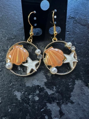 Runda örhängen med snäcka och stjärna - Unika örhängen i guldfärgad metall med rund form, dekorerade med orange snäcka, vit sjöstjärna och små pärlor. Perfekta för dig som gillar strandvibbar och detaljerade accessoarer. Krokar i guldfärg för enkel användning.