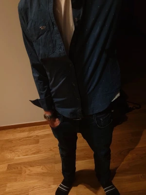 Mörkblå skjorta från Hollister XS - Säljer en mörkblå skjorta från Hollister i stretchigt bomullsmaterial. Skjortan har långa ärmar, knappar framtill och en liten logga på bröstet. Perfekt för dig som gillar en klassisk men avslappnad stil.