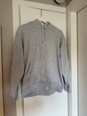 Ljusgrå hoodie med ficka och huva - En basic ljusgrå hoodie med huva och dragsko, samt en stor magficka framtill. Liten fläck på snöret.