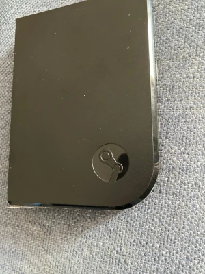 Steam Link  - Steam Link är en svart, rektangulär spelstreaming-enhet i plast med rundade hörn och Steam-loggan på framsidan. Perfekt för att streama spel från din PC till TV:n och få en smidig gamingupplevelse i vardagsrummet.