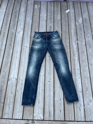 Nudie Jeans Big Bengt  - (Jeansen är discountinued) Klassisk modell Big Bengt från Nudie Jeans Co i blå denim, med en blekt och sliten tvätt samt naturliga veck och slitagemönster. Dessa jeans med rak passform har signatursömmar i kontrasterande orange färg, gylf med knappar och den ikoniska Nudie Jeans-vågen på bakfickorna. W29 L32  #nudiejeans #bigbengt #denim #jeans #straightleg #fadeddenim #distressedjeans #mensfashion #secondhand #preloved #sustainablefashion #bluejeans #buttonfly #designerjeans #casualwear