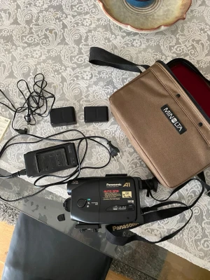 Vintage Panasonic VHS-C Videokamera (Y2K / Retro) 🎥📸 - Säljer ett supermysigt retro camcorder-kit med en Panasonic NV-A1 från 90-talet! Perfekt för att få den där äkta, gryniga lo-fi/skate-estetiken på dina videos som inte går att få med en smartphone.   Hela kittet säljs tillsammans och du får med: - Kameran (Panasonic NV-A1) med linsskydd. - Original nätadapter/laddare med alla sladdar (går att driva direkt via vägguttaget!). - En snygg, brun Minolta-väska med axelrem. 