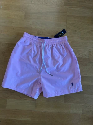 Rosa badbyxor från Polo Ralph Lauren - Snygga ljusrosa badbyxor från Polo Ralph Lauren med klassisk marinblå logga broderad på benet. Modellen har elastisk midja med vit dragsko och sidofickor. Perfekta för stranden eller poolen, tillverkade i snabbtorkande material.
