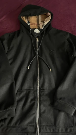 Svart hoodie från Burberry med dragkedja - Svart hoodie från Burberry med dragkedja och klassiskt rutigt foder i huvan. Hoodien har dragsko, fickor framtill och en diskret broderad logga på bröstet. Perfekt för en stilren och avslappnad look.