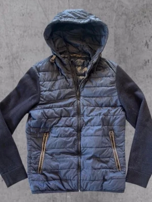 Massimo Dutti cardigan - Snygg blå pufferjacka med vadderad kropp och stickade ärmar. Jackan har dragkedja framtill, två fickor med dragkedja och en fast huva. Perfekt för kalla vinterdagar och ger en sportig look.