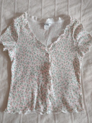 Blommig ribbad topp från Monki med vågig kant och knäppning - Supersöt ribbad topp från Monki med blommigt mönster i rosa och grönt på vit botten. Toppen har v-ringning, korta ärmar, knäppning i fram och volangkant längs kanterna 🤍 Storlek XS. Fint skick. 