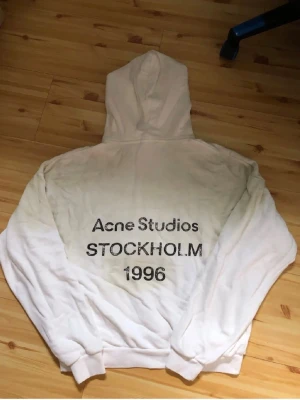 Vit hoodie Acne Studios Stockholm - Säljer en vit hoodie från Acne Studios med trycket 'Acne Studios STOCKHOLM 1996' på ryggen. Hoodien har klassisk huva och ribbade muddar. Perfekt för dig som gillar streetwear och vill ha en clean look med en touch av Stockholm.