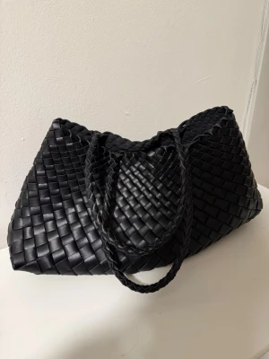 Svart flätad handväska i skinnimitation - Säljer en svart handväska med flätad design och dubbla handtag. Väskan har en mjuk, rymlig form och är tillverkad i skinnimitation. Perfekt för dig som gillar trendiga accessoarer med en unik twist.