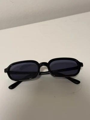 Svarta rektangulära solglasögon Bik Bok  - Snygga svarta solglasögon med rektangulära bågar och mörka glas. Klassisk och stilren design som passar till alla outfits. Bågarna är i plast och glasen är mörkt tonade för ett coolt uttryck.