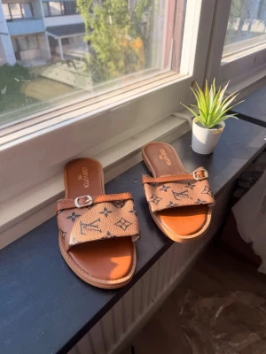 Loui Vuitton sandaler - Säljer ett par bruna sandaler med klassiskt likt  monogrammönster och guldfärgat spänne. Sandalerna har öppen tå och platt sula, tillverkade i konstläder med en bred rem över foten. Perfekta för en trendig och avslappnad look.