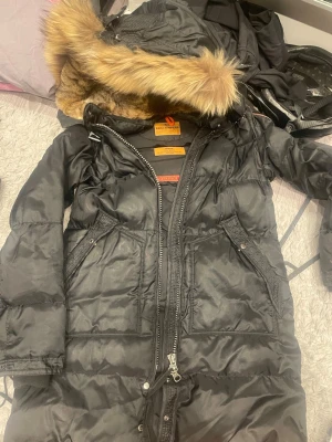 Svart lång dunjacka från Parajumpers - Säljer en svart lång dunjacka från Parajumpers Masterpiece Series. Jackan har stor huva med avtagbar fuskpäls, dragkedja framtill och två stora fickor med lock. Perfekt för kalla vinterdagar och riktigt snygg streetstil.