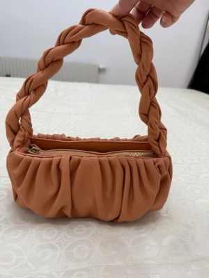 Aprikos handväska med flätat handtag - Trendig handväska i mjukt, aprikos konstläder med rynkad design och ett rejält flätat handtag. Väskan har guldfärgad dragkedja och ett rymligt huvudfack. Perfekt accessoar för att lyfta din outfit.