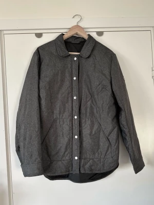 J Lindeberg- Mörkgrå quiltad jacka - storlek 44 / XL - Tidlös grå J.Lindeberg jacka / täckjacka i storlek 44 med diskret stilrent quiltat mönster. Klassisk modell med rak krage, silverfärgad knäppning och fickor framtill. Praktiska tryckknappar i midjan baktill för justering av passformen. Diskret logo på vänster arm och på tryckknappar. Dubbelgängad dragkedja, så man kan öppna jackan både uppifrån och nedifrån. Nypris 1800:-. Mycket gott skick, som ny!