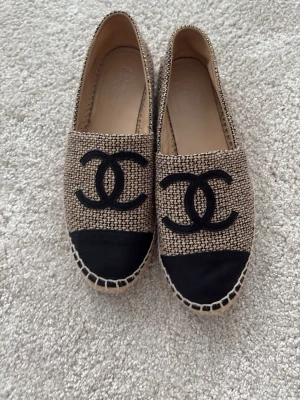 Beige och svarta loafers med logga från Chanel - Säljer ett par snygga loafers i beige och svart med tydlig logga framtill. Skorna har en flätad sula och ovandel i textil med svart tåparti. Perfekta för dig som gillar stilrena detaljer och vill ha något bekvämt men ändå trendigt.