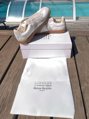 Maison Margiela vita sneakers - Maison Margiela sneakers i vitt läder och mocka med beige detaljer och klassisk gummisula. Låg modell med stilren design och diskret logga på plösen. Perfekta för dig som gillar minimalistisk och trendig stil.