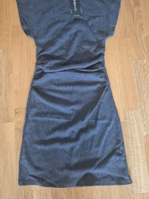 Mörkgrå  klänning - Snygg mörkgrå klänning i kort modell. Klänningen har en figurnära passform och är tillverkad i ett mjukt, stretchigt material som ger en bekväm känsla. Perfekt för dig som vill ha en enkel men stilren look.