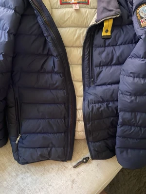 Mörkblå dunjacka från Parajumpers - Snygg mörkblå dunjacka från Parajumpers med quiltad design och gul detalj vid halsen. Jackan har två dragkedjeförsedda fickor framtill och patch på ärmen. Insidan är ljus och jackan har hög krage. Perfekt för kalla vinterdagar.