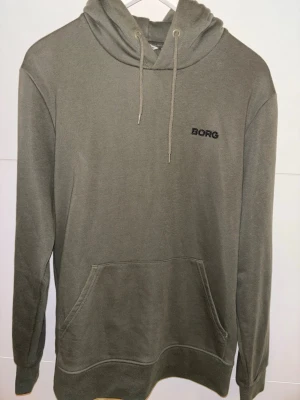 Olivgrön hoodie från Björn Borg - Snygg olivgrön hoodie från Björn Borg med broderad logga på bröstet. Klassisk känguruficka framtill, dragsko i huvan och mjukt material som känns skönt mot huden. Perfekt för en avslappnad och stilren look.