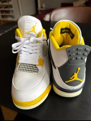 Air Jordan 4 Retro White/Yellow - Snygga Air Jordan 4 Retro sneakers i vitt läder med gula och grå detaljer. Skorna har klassisk mesh på sidan, gula accenter på sulan och innersidan samt Jumpman-logga på plös och häl. Perfekt för dig som vill sticka ut med färg och ikonisk design.