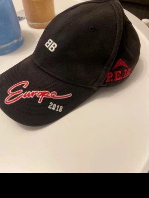 Svart Balenciaga keps - Säljer en svart keps från Balenciaga med broderad text 'Europe 2018' i rött och vitt framtill samt 'P.E.18' på sidan. Klassisk böjd skärm och justerbar passform. Unik och extremt efterfrågad modell. Box och dustbag ingår 