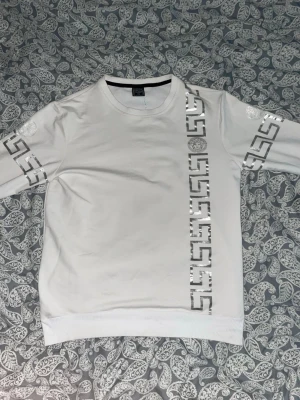  sweatshirt vit med silver mönster Strl S - Snygg vit sweatshirt med silverfärgat tryck i grekiskt mönster längs ärmarna och framtill samt dekorativ logga på bröstet. Bekväm passform och mjukt material – perfekt till vardags eller streetwear-outfits.  Använd men i fint skick, inga större skador Färg: Vit med silvertryck.                                             storlek: S Passform: Normal Perfekt att matcha med jeans, mjukisbyxor eller shorts.  Kategori: Sweatshirt / Tröja Skick: mycket bra skick