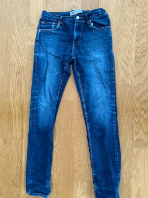 Blå skinny jeans från Levi's - Säljer ett par klassiska blå slim fitt jeans från Levi's med fem fickor och snygga kontrastsömmar. Jeansen har en normalhög midja och är tillverkade i stretchigt denim för skön passform. Perfekta till en avslappnad stil. Storlek 176, resår midja.