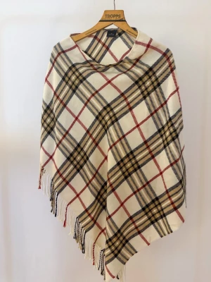 Rutig ponchosjal med fransar - Stilren ponchosjal i vitt med rutigt mönster i svart, beige och rött. Sjalen har fransar längs nederkanten och är tillverkad i ett mjukt tyg som ger en skön känsla. Perfekt för lager-på-lager och att hålla värmen under kyliga dagar.