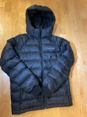 Svart dunjacka från Peak Performance - Snygg svart dunjacka från Peak Performance med huva och dragkedja framtill. Jackan har diskret logga på bröstet och är vadderad för extra värme. Perfekt för kalla vinterdagar och har en stilren, sportig look. Köpt för 2 månader sedan inga tecken på användning, nypris 2000kr.