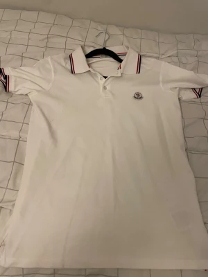 Vit Moncler pikétröja med rödblå detaljer - Klassisk vit pikétröja från Moncler med rödblå ränder på krage och ärmslut. Tröjan har knappslå framtill och Moncler-logga broderad på bröstet. Perfekt för en clean och stilren look. Priset kan diskuteras 